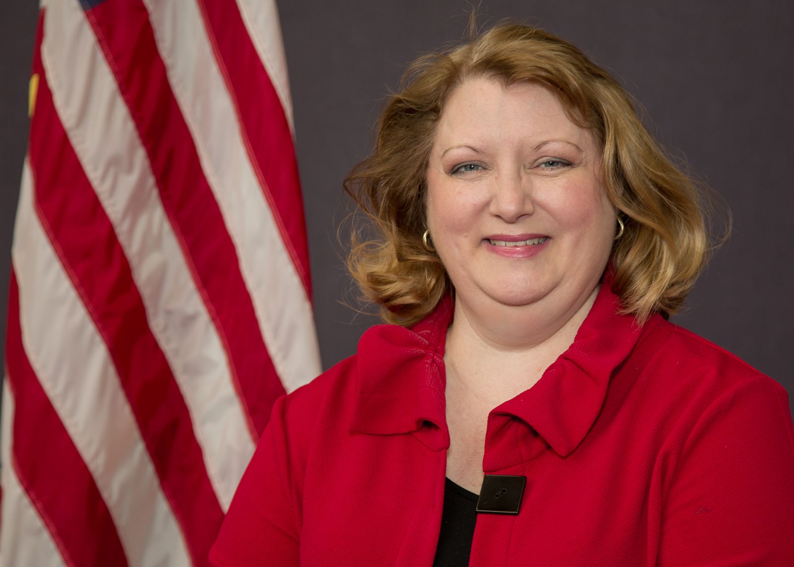 A blonde, white caucasian, Gena Sapp, is wearing a bright red collared jacket over a black shirt and is pictured with the United States flag standing in the back left against a grey background.