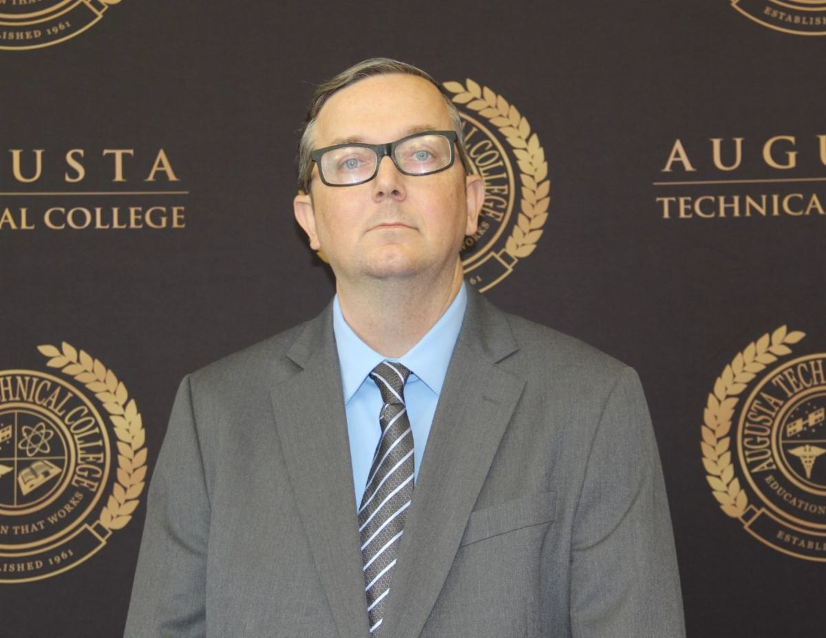 Caucasian male wearing black glasses, gray suit jacket, blue collared shirt, gray and white striped tie, standing against a black background with the words Augusta Technical College in gold