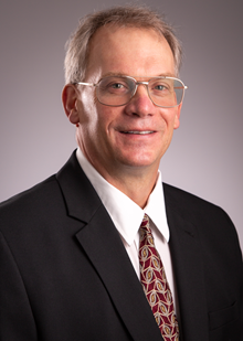 Caucasian male, smiling, wearing glasses, whit collared shirt, black suit jacket, red tie with design