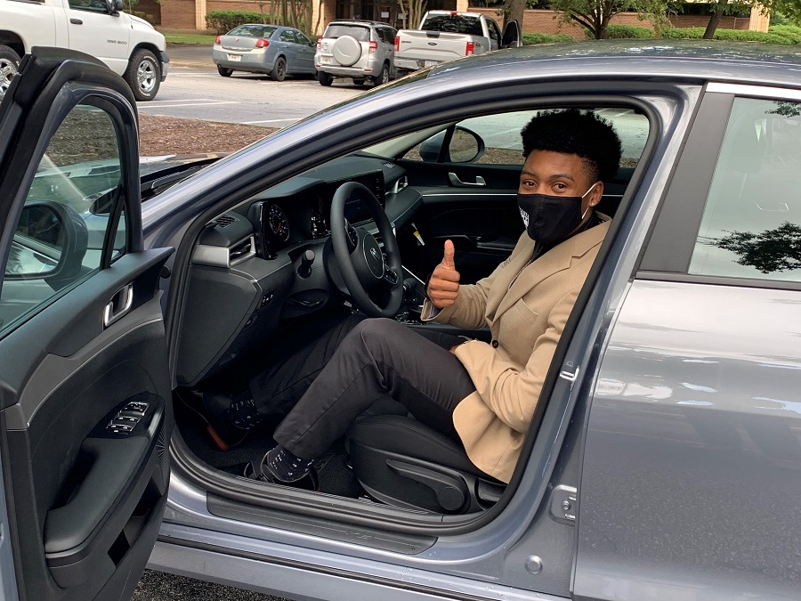 2020 state GOAL winner, Rah'Mere Williams wearing a black Augusta Technical College mask, a tan suit jacket and grey pants, sits in his new silver 2021 KIA K5 with the driver side door open and posing with a thumbs up for the camera.