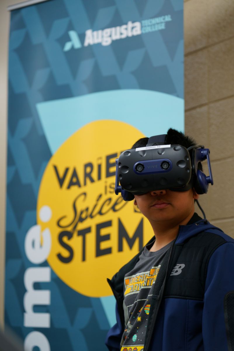 An African American male stands solemnly wearing VR equipment in front of an Augusta Technical College VARIETY is Spice STEM banner.