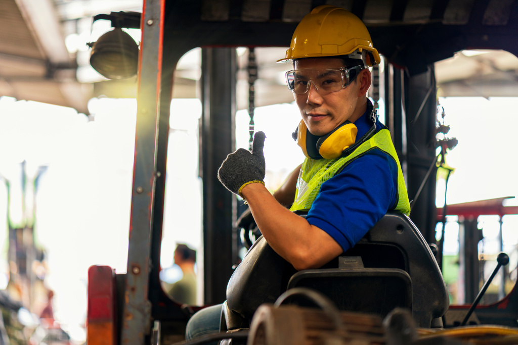 A forklift operator sits in the seat of a forklift wearing a yellow hard hat, safety glasses and yellow safety vest with yellow noise cancelling headphones around his neck.