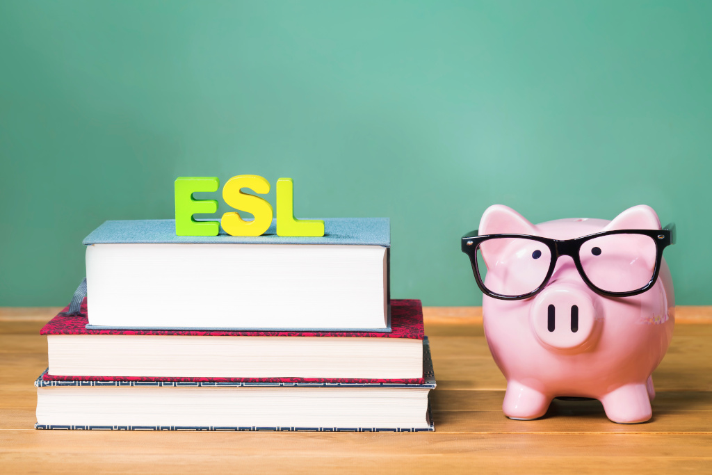 Three books of various size and thickness are stacked on a wooden desk with the letters ESL in yellow and lime green sitting on top against a chalkboard green background. A pink ceramic pig wearing black frame glasses sits to the right of the books.