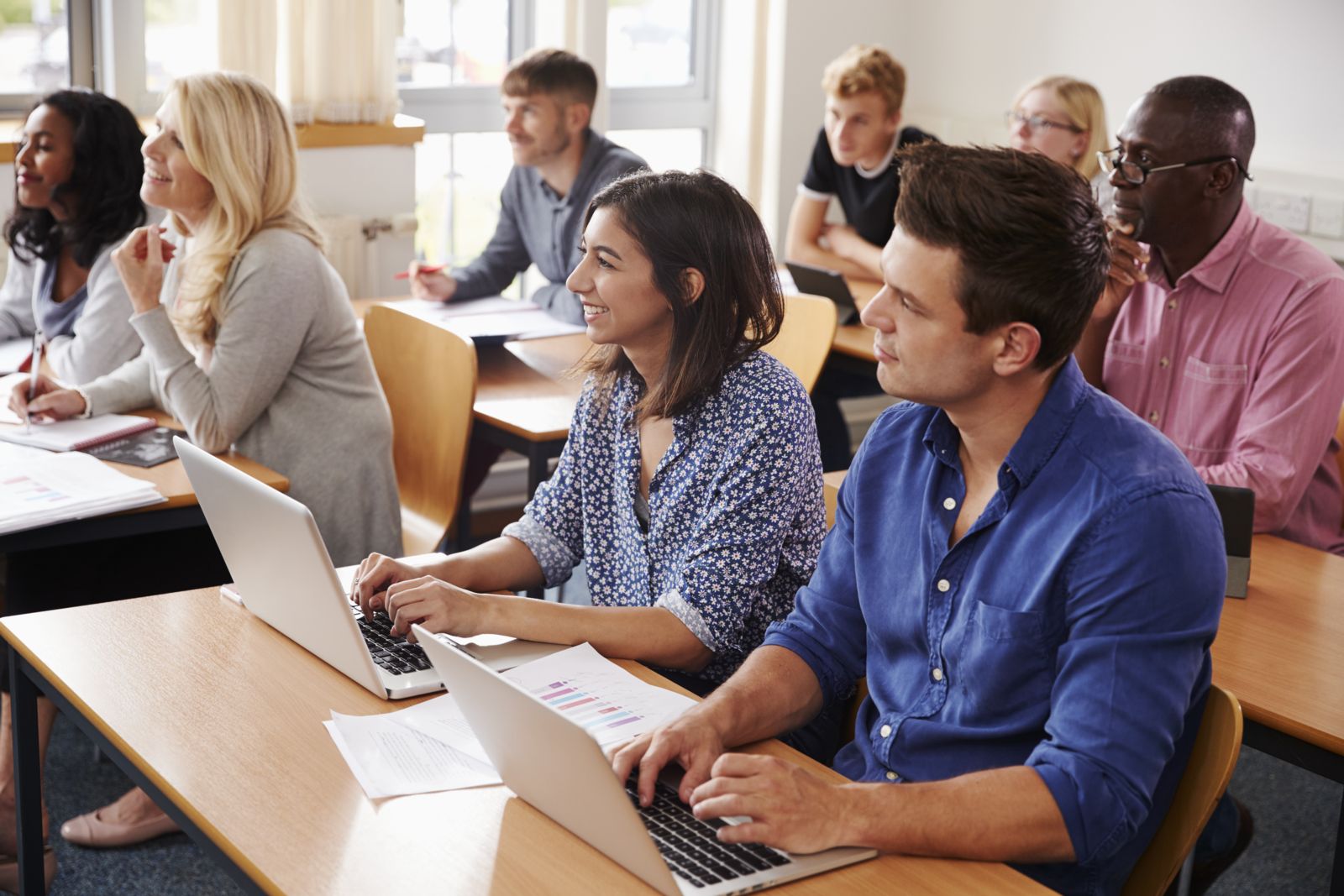 A mixed group of adults, sitting at desks in a classroom and directing their attention to the front of the sunny classroom. Some are taking notes on laptops, others on paper, and others are not taking notes.