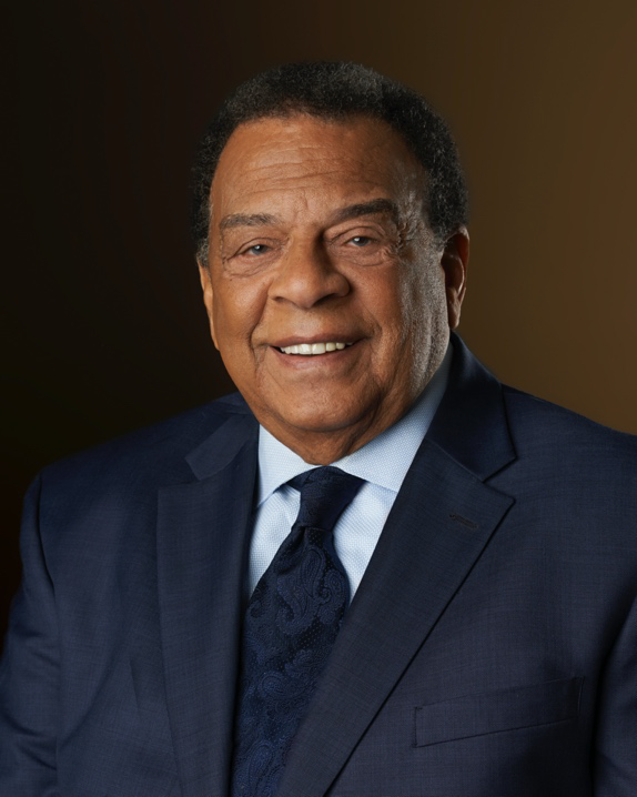 Ambassador Andrew Young, an older African American Male, smiles wearing a navy suit with a light blue collared shirt and a navy blue tie with a floral pattern.