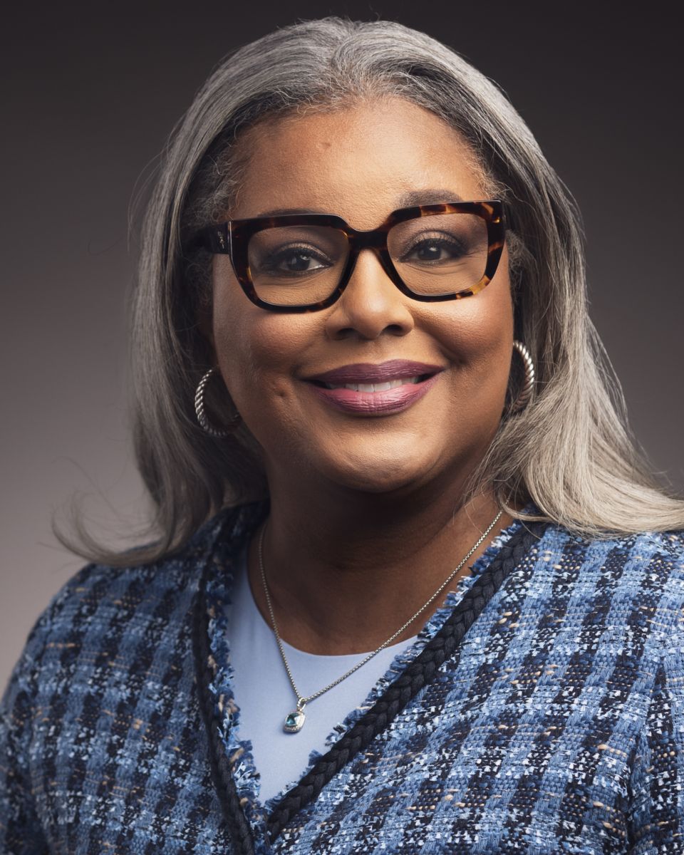 Ms. Collette D'Antignac, an African American female with long grey hair, smiles at the camera wearing brown rimmed glasses and a blue dress shirt with a blue plaid cardigan.