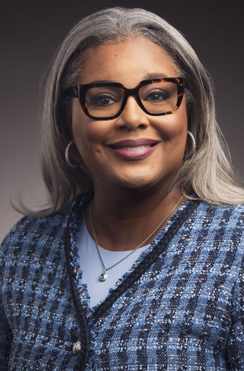 Collette D'Antignac, an African American female, smiles at the camera wearing brown rimmed glasses and a solid blue shirt under a blue plaid sweater.
