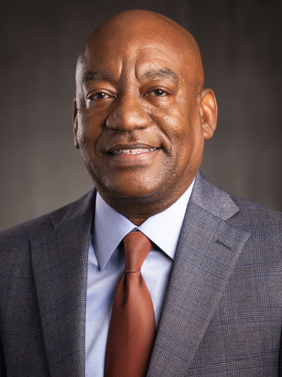 Mr. Derric Terrentine, an African American male, smiles wearing a light blue collared shirt and burnt orange tie with a light grey suit jacket on a grey background.