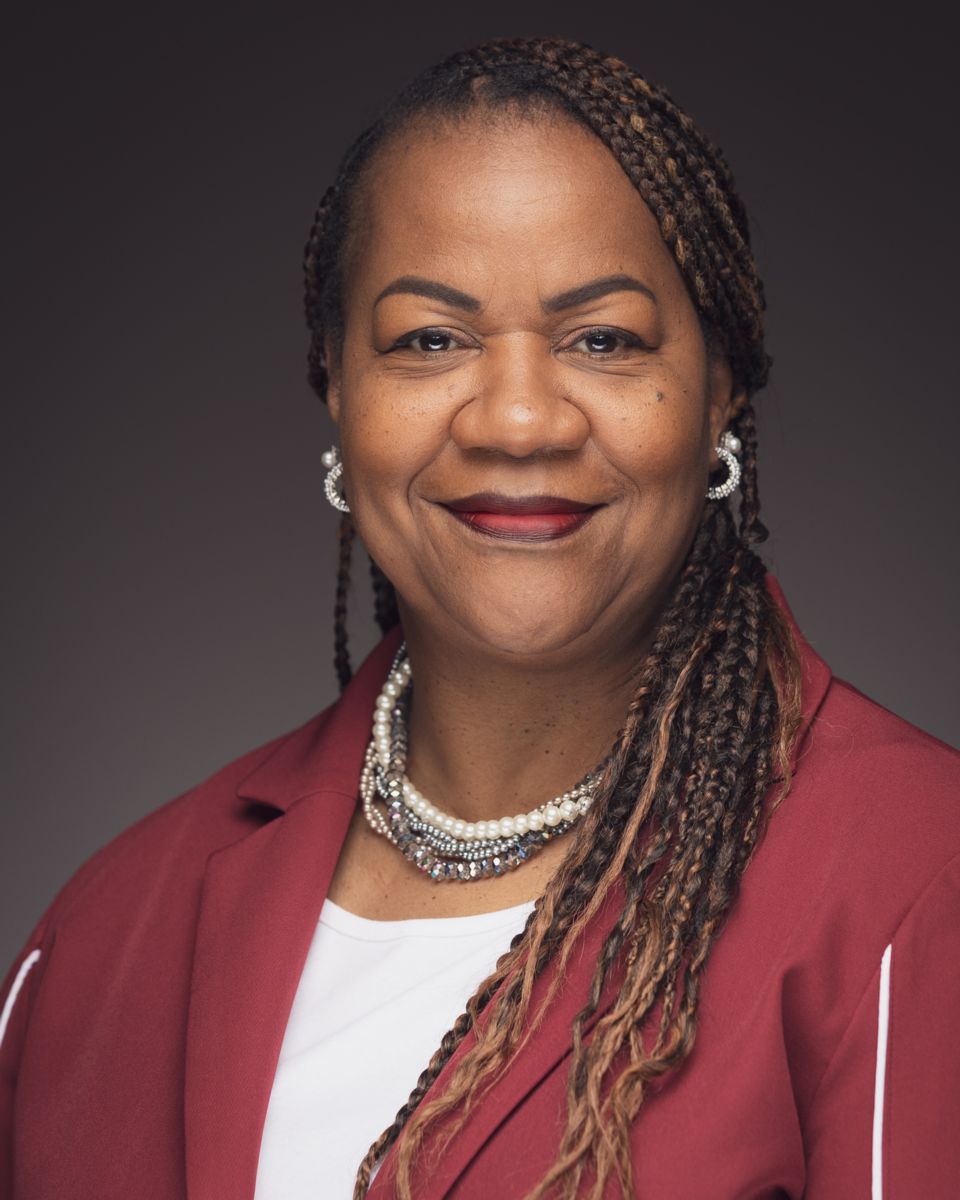 Professional headshot of Grace Scott smiling at the camera, with long highlighted braids swept over one shoulder, wearing decorative earrings, layered pearl and beaded necklaces, a white top, and a burgundy blazer, against a dark gray studio background