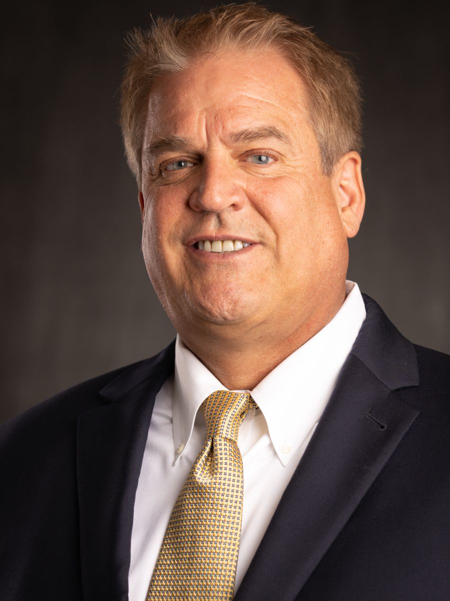 Mr. Jon Dollan, a Caucasian male with short grey and brown hair smile wearing a white collared shirt, gold tie with white dots, and a black suit jacket in front of a grey background.