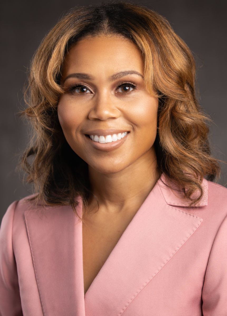 Ms. Katrell Nash, an African American female with long caramel colored hair, smiles wearing a pink suit jacket on a grey background.