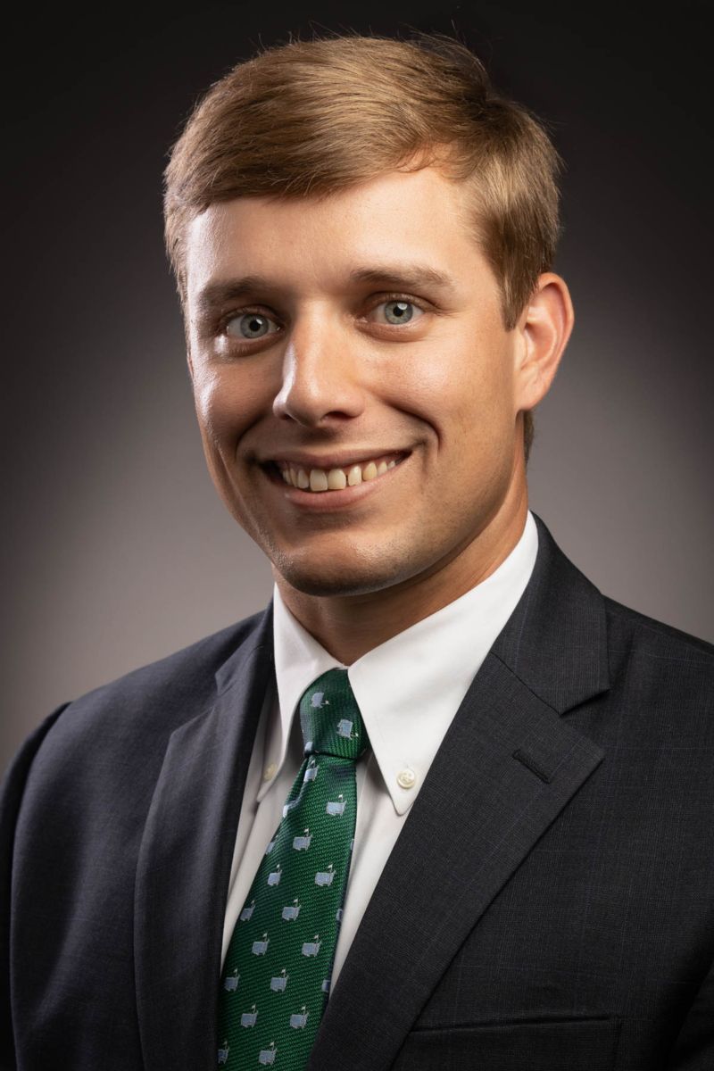 Mr. Nicholas Wood, a Caucasian male, smiles wearing a black suit with a white button down shirt and a green tie decorated with white apple symbols.