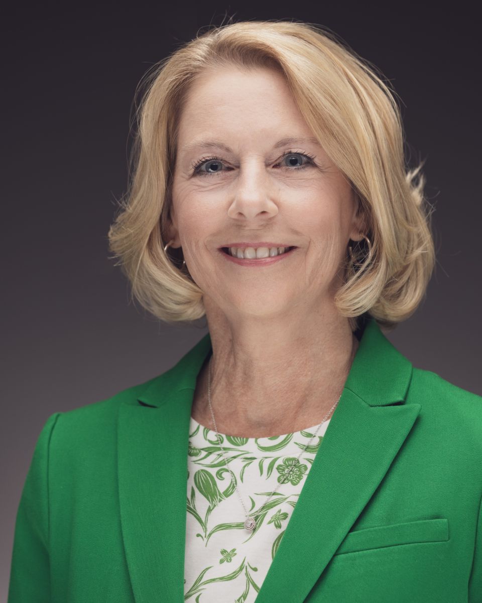 Professional headshot of Tammy Shepherd smiling at the camera, with shoulder-length blonde hair, wearing a bright green blazer over a white top with a green floral pattern and a silver necklace, against a dark gray studio background