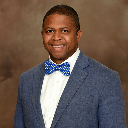 Mr. Donovan Lee-Sin, an African American male, smiles at the camera wearing a light blue suit with a white collared shirt and blue and white striped bow tie.