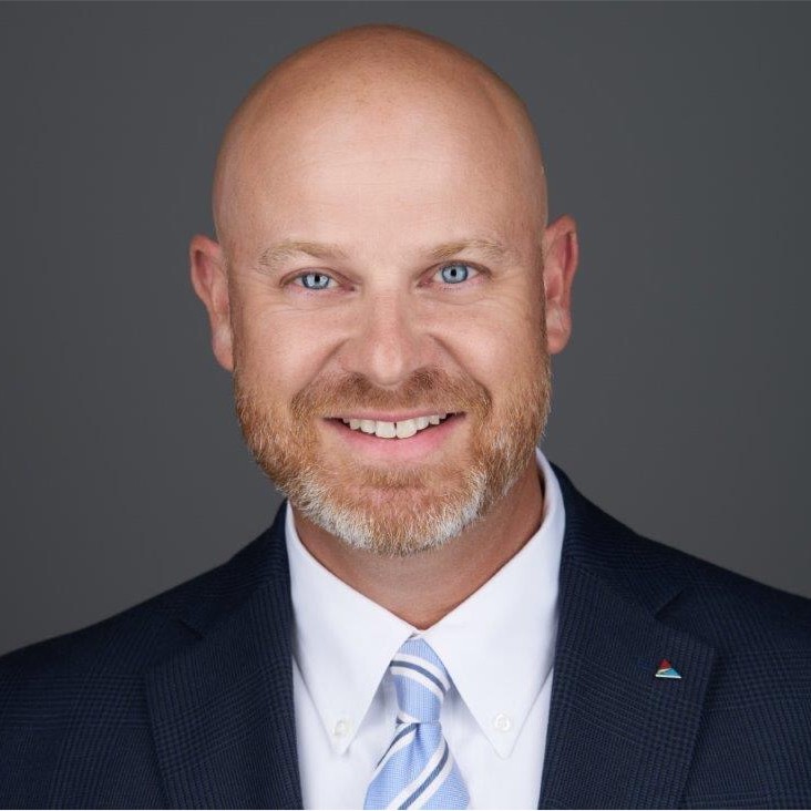 Mr. Kerry Bridges, a Caucasian male with a red and gray beard, smiles at the camera wearing a navy blue suit and is pictured against a grey background.