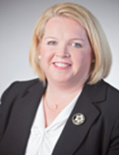 Ms. Reyne Gallup a Caucasian female with short blonde hair smiles wearing a black suit jacket with a white collared shirt.