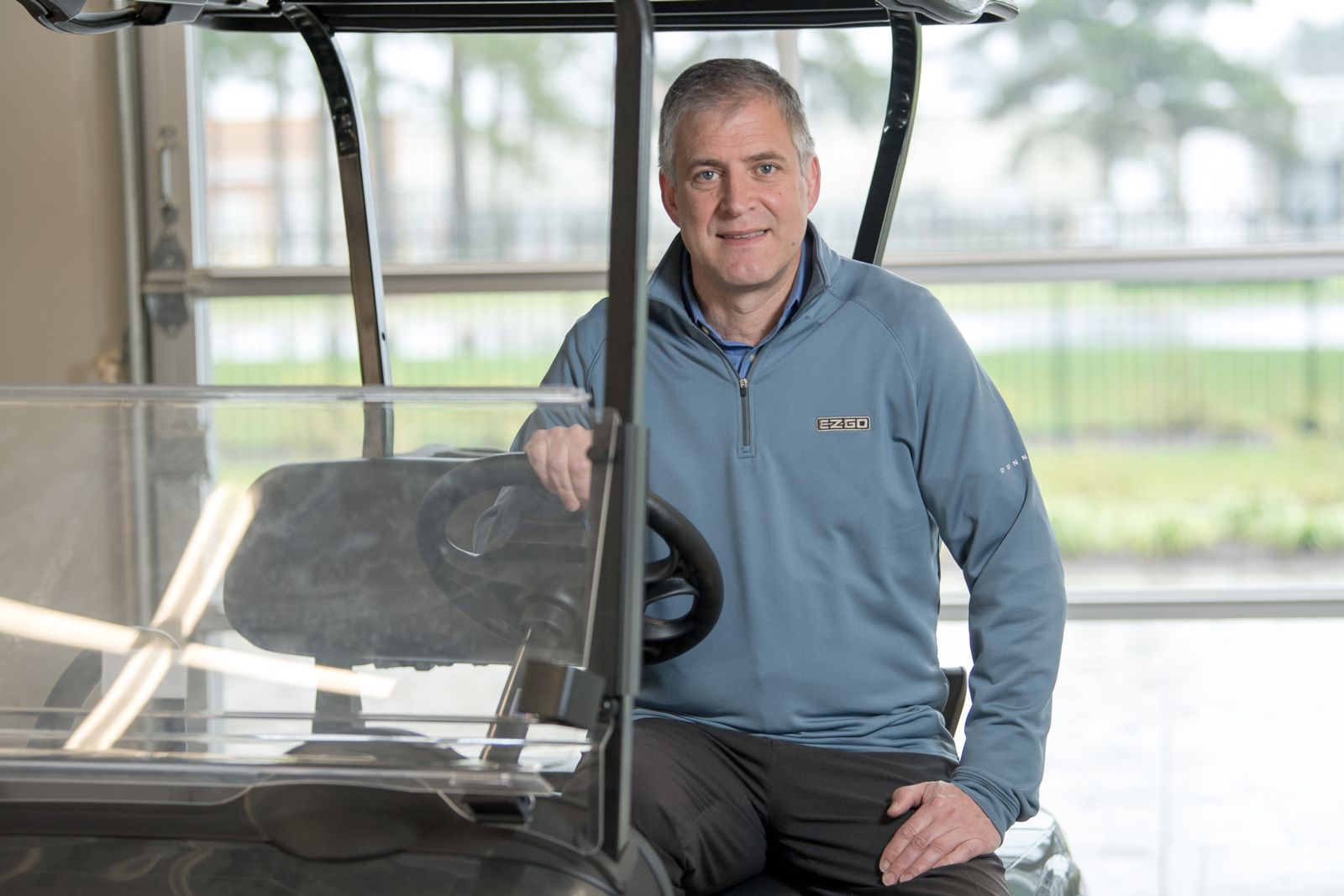 Mr. Gunnar Kleveland, a Caucasian male, sits at the wheel of a golf cart wearing a blue fleece pullover and grey slacks while smiling at the camera.