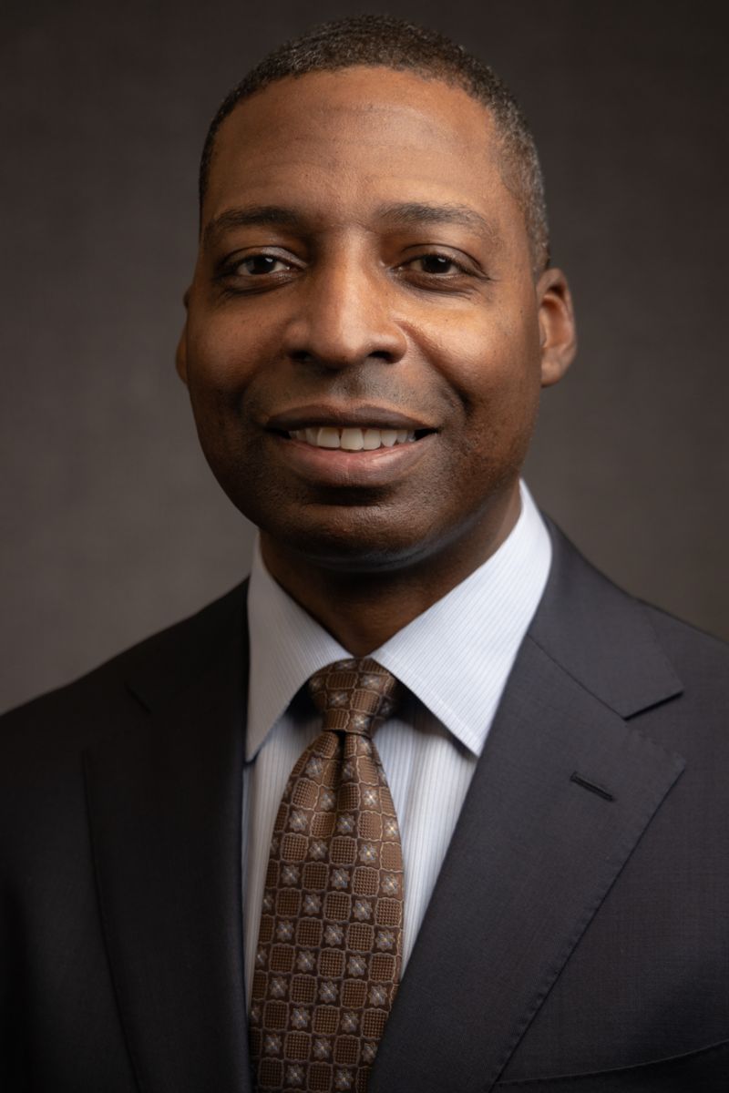 Mr. James Heffner, an African American male, wears a charcoal gray suit with a brown and tan patterned tie and is pictured against a gray background.