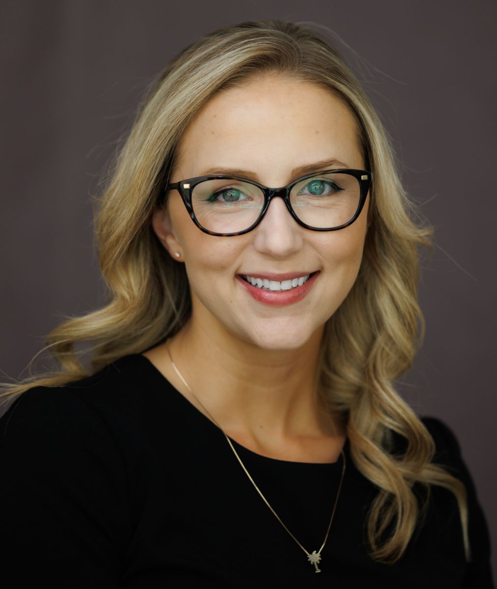 Ms. Marissa Smith, a Caucasian female with long blond hair wears black rimmed glasses and a black suit jacket with a white shirt while smiling at the camera.