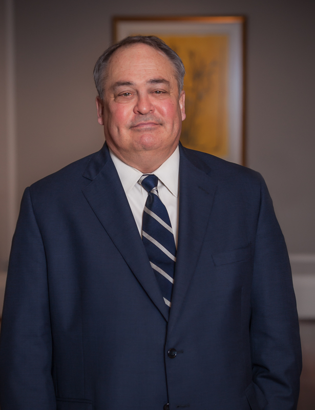 Caucasian male, wearing a blue suit with buttons, white collared shirt, blue and silver striped tie, standing against a neutral-colored background with a painting on the wall, posing for a photo.