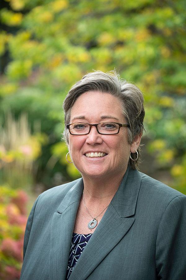 Dr. McElroy Hooper, a Caucasian female with short blonde hair, smile wearing a grey suit jacket and glasses.