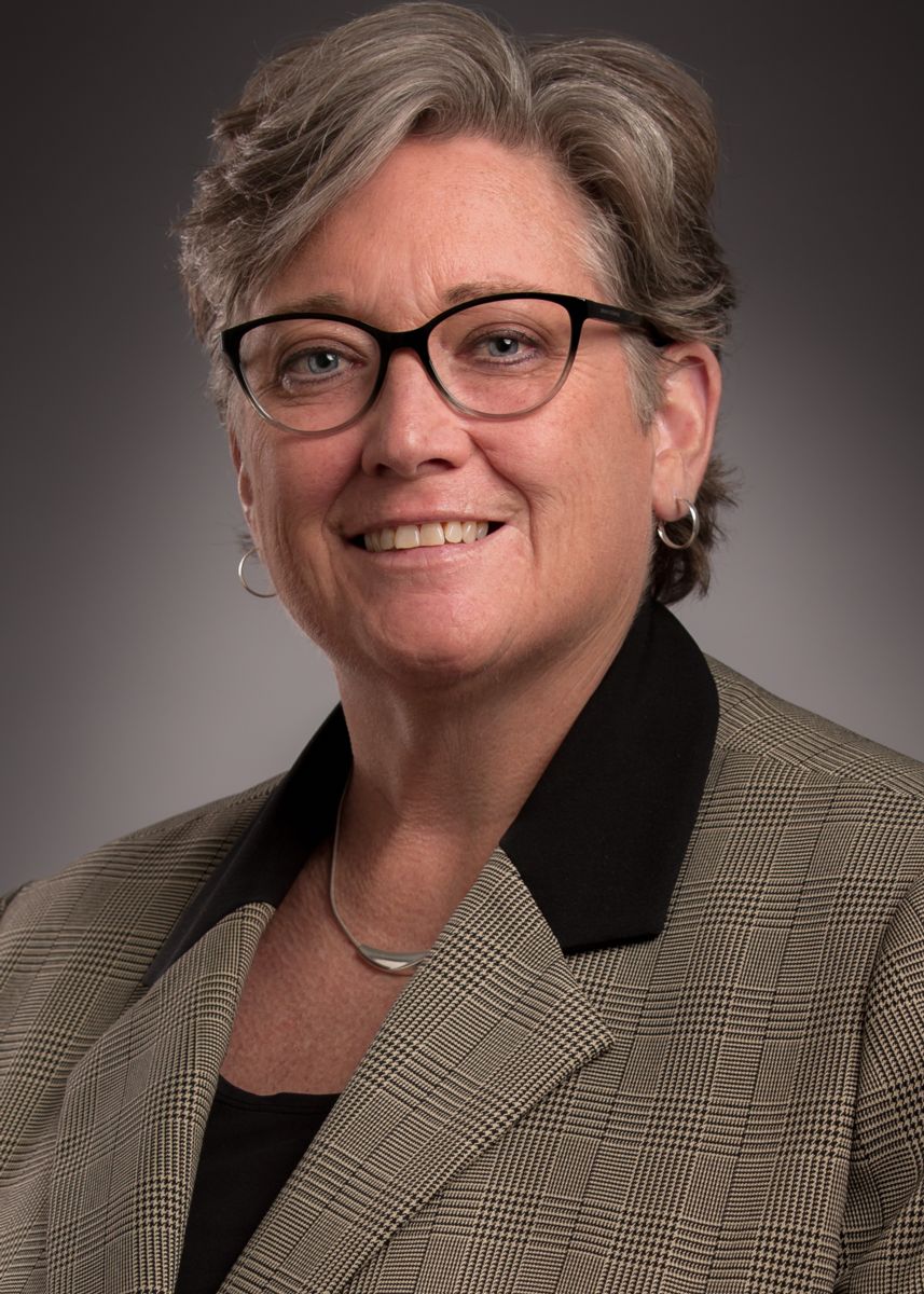 Dean Kellie McElroy Hooper, a Caucasian female with short grey and brown hair, smiles wearing glasses, a brown suit jacket with a black collar, and a black shirt.