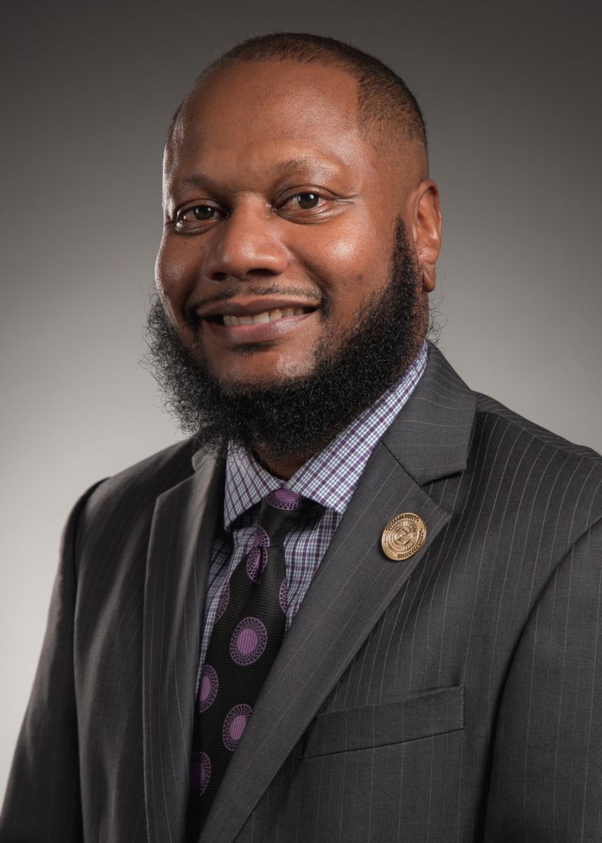Dean Quentin Cooks smiles at the camera wearing a silver striped grey suit with a blue striped white shirt  and a dark purple tie with lighter circles on it.