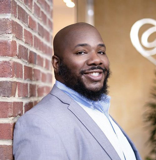 Mr. Shaun Andrews, an African American male, smiles at the camera while leaning against a brick wall wearing a silver suit with a grey and white striped collared shirt.