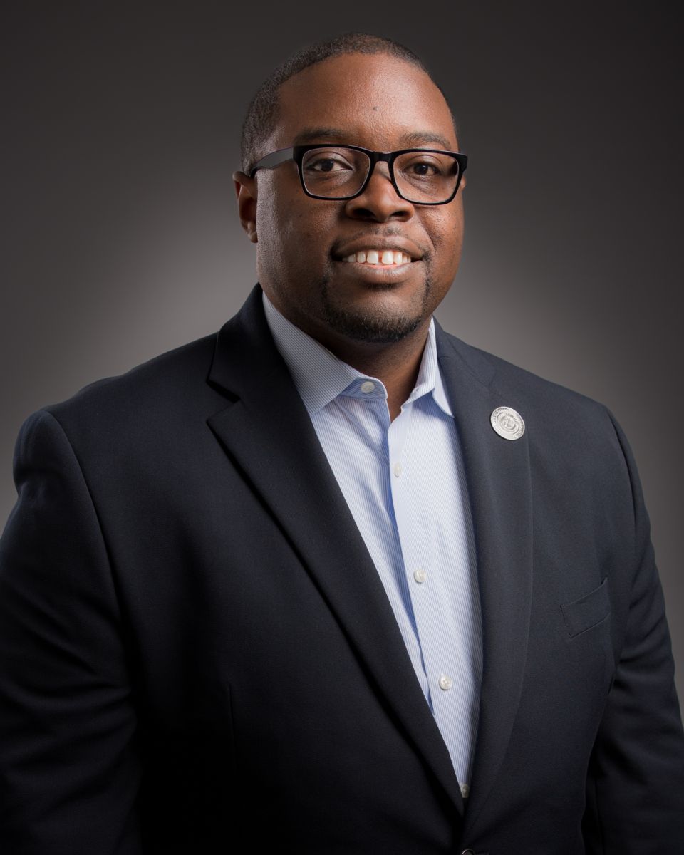 Dr. Jermaine Whirl, a smiling African American male, wearing a black suit with a white collared shirt, and black rimmed glasses. A round gold pin is on his left lapel.