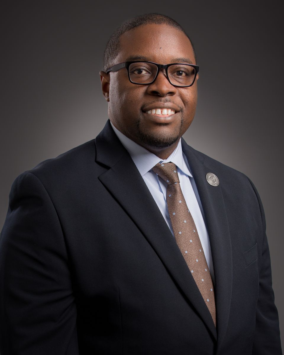 Dr. Jermaine Whirl, a smiling African American male, wearing a black suit with a white collared shirt, a brown tie with gold dots, and black rimmed glasses. A round gold pin is on his left lapel.
