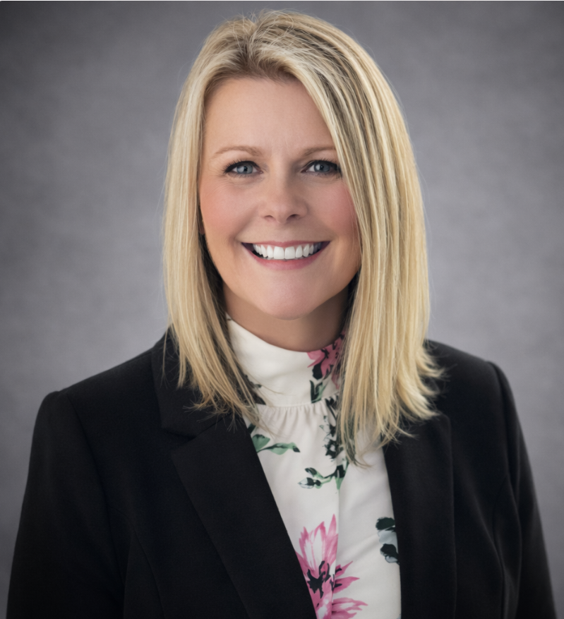 An Caucasian female, wearing a black suit jacket white blouse with a pattern and a gray background