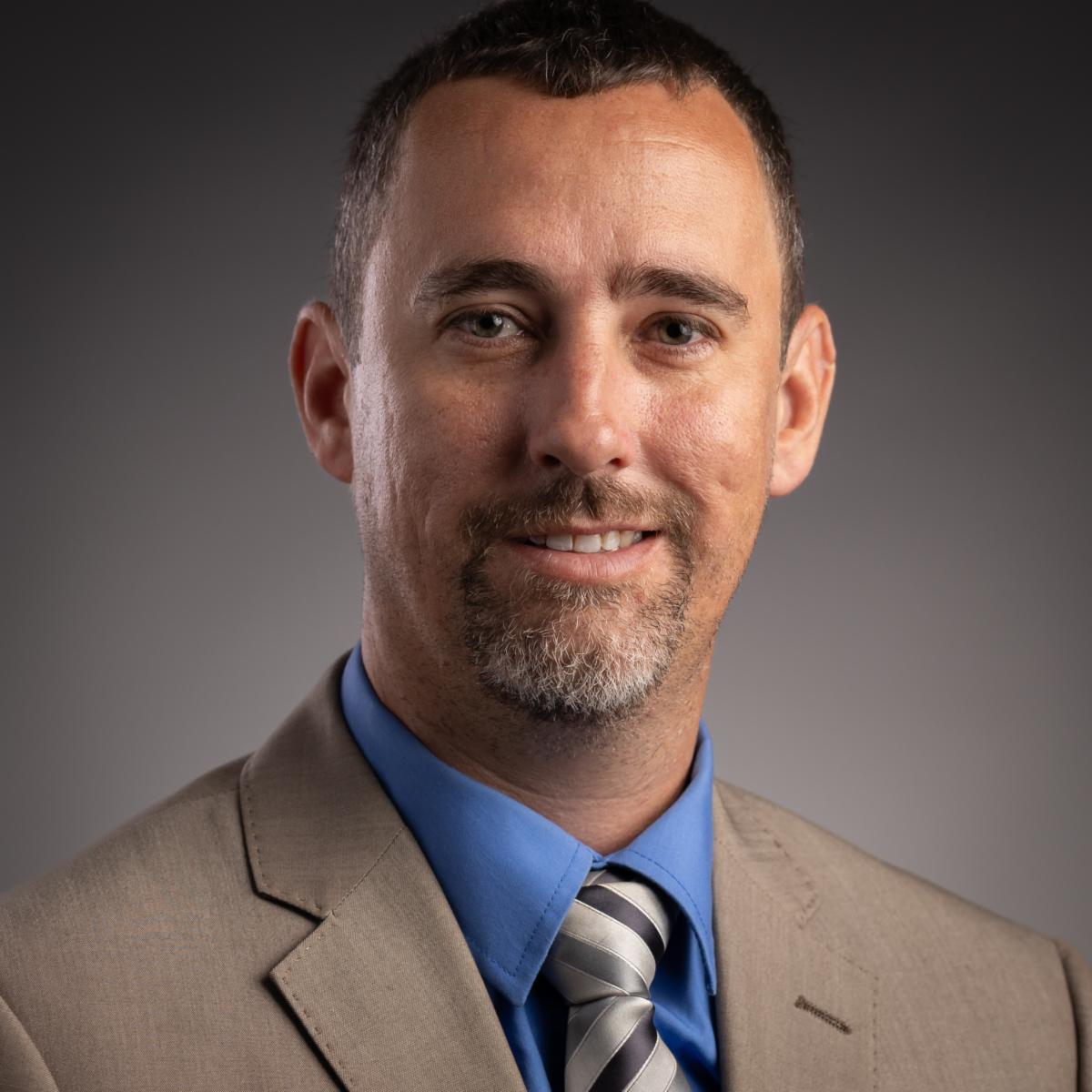 Mr. Thomas Hippchen, a Caucasian male, smiles at the camera wearing a tan suit with a blue collared shirt and a tan and brown striped tie.