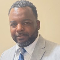 An African American male, wearing a gray suit with a tan background.