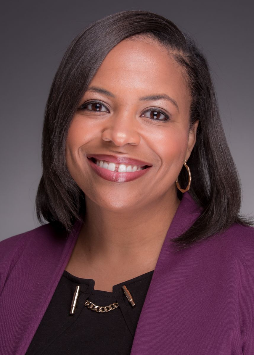 Ms. Christine Ball, an African American female with shoulder length dark brown hair, smiles at the camera wearing a purple suit jacket and a black dress shirt with a bronze metal clasp on the front and gold hoop earrings.