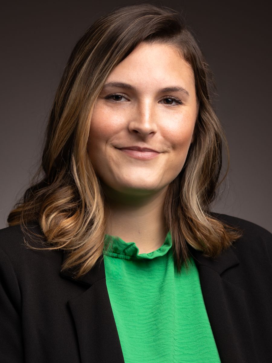 Ms. Gabriella Nanes, a Caucasian female, smiles at the camera wearing a green shirt with a black dress jacket.