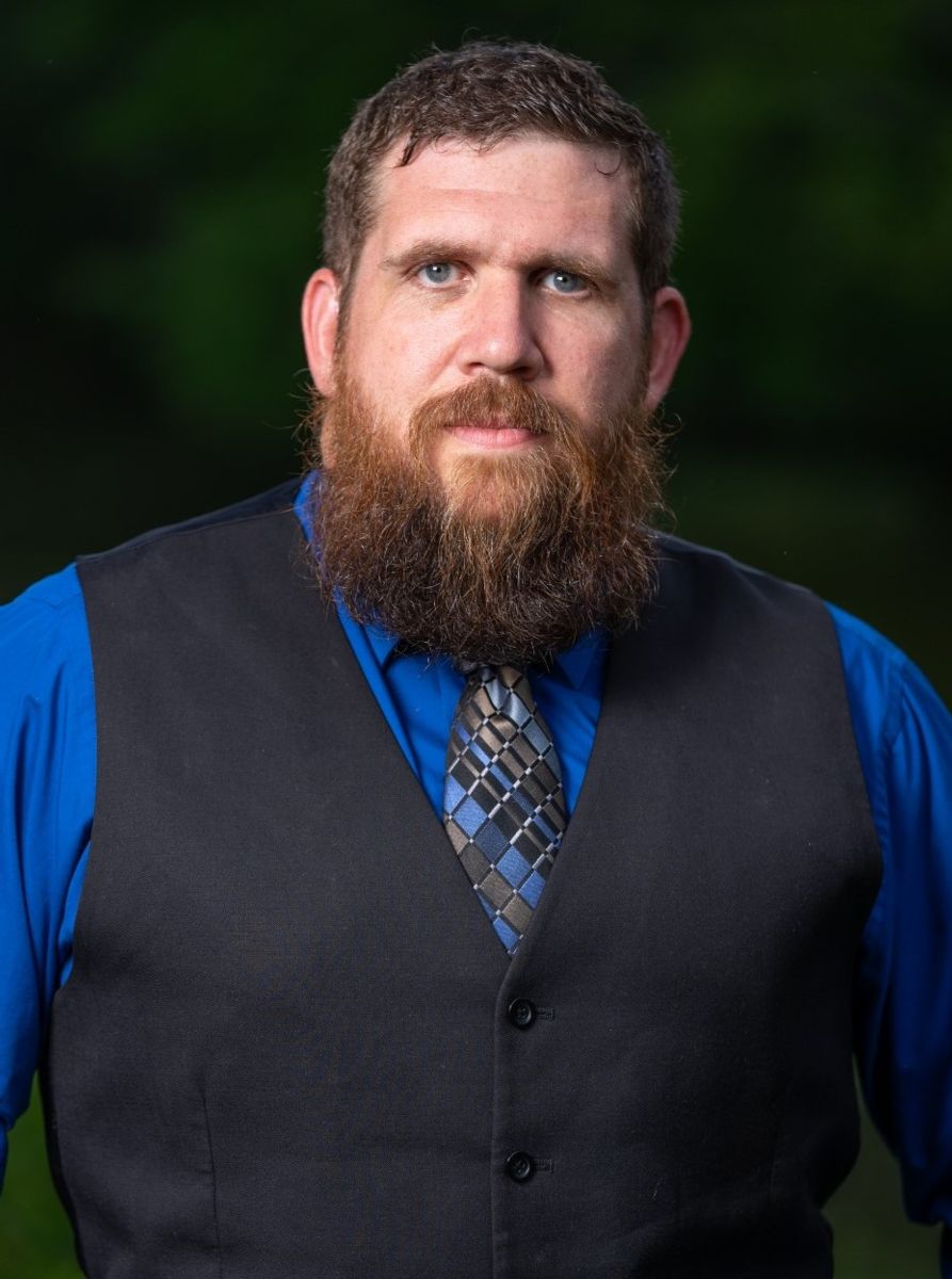 A Caucasian male with brown hair, brown beard, wearing a blue dress shirt and vest with tie with a light blue gold black and blue checkered pattern.