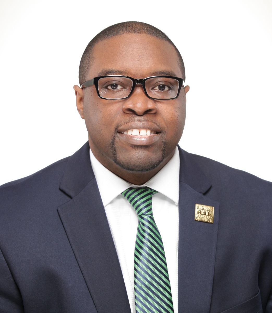 Smiling, African American man wearing a blue suite, green and blue striped tie, white collared shirt, gold lapel pin posing for a photo against a white and gray background
