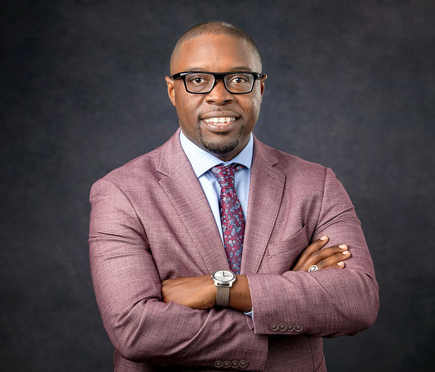 Dr. Jermaine Whirl smiles at the camera wearing a red suit and light blue collared shirt with a red and blue patterned tie pictured against a grey background.