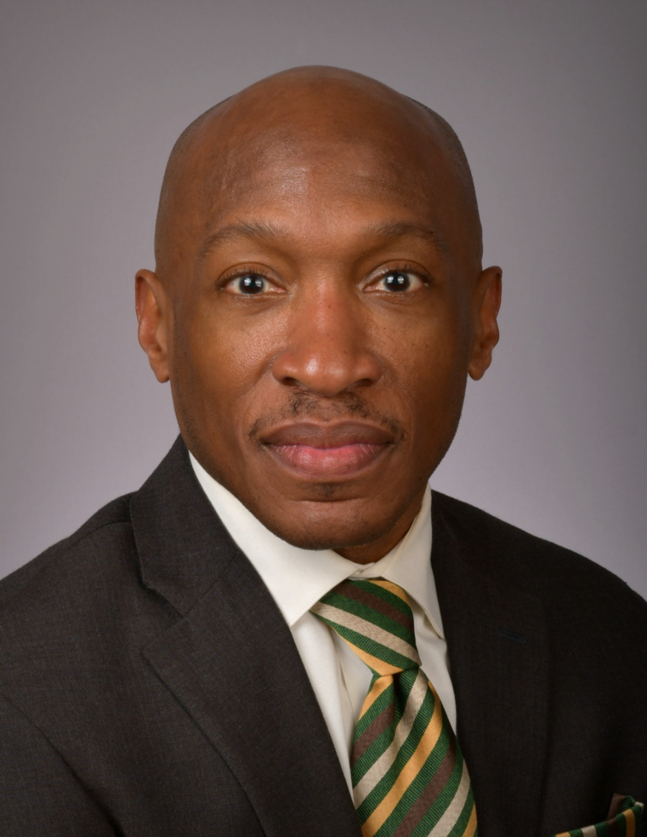 Dr. Kendricks Hooker, an African American male, is pictured wearing a black suit jacket with a white collared shirt and green, white and yellow striped tie against a light grey background.