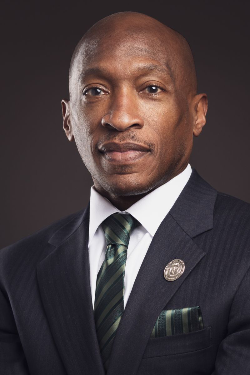 Dr. Kendricks Hooker , an African American male, wears a suit with a black jacket with a green tie, brownish background