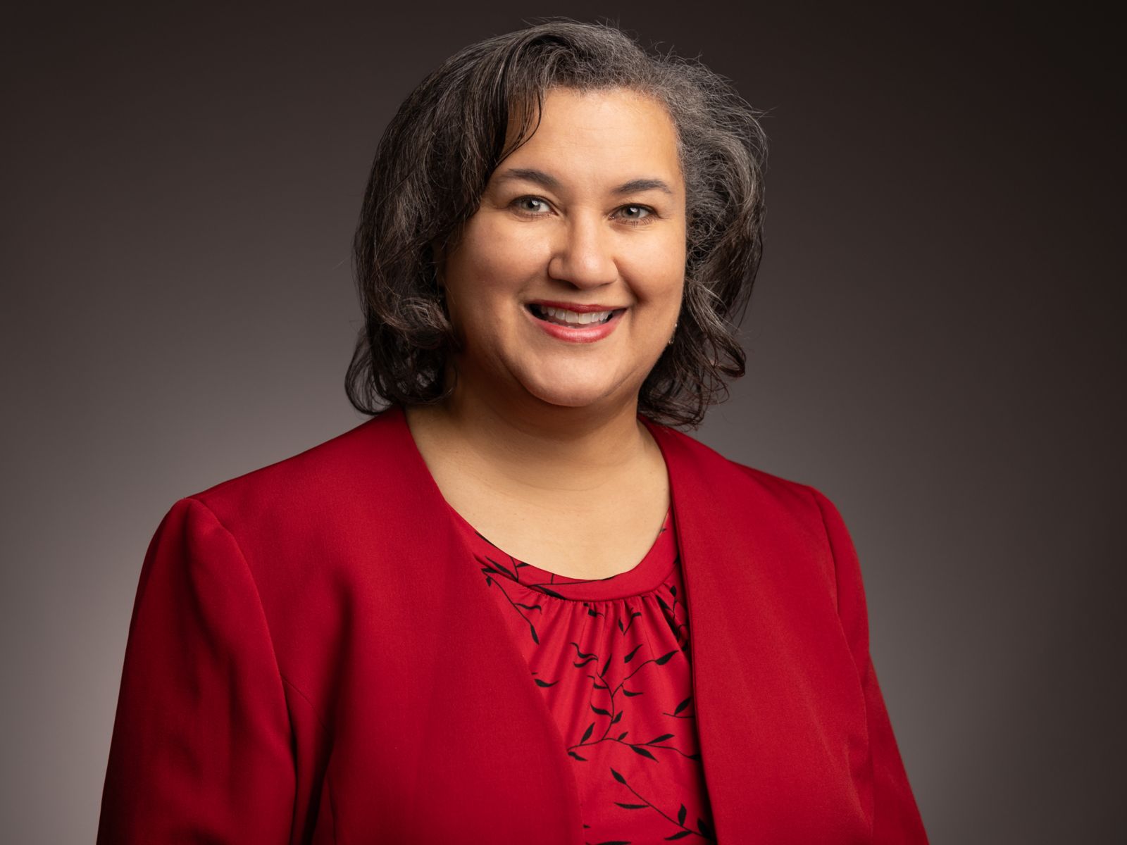 Dr. Melissa Frank-Alston, a Caucasian female with shoulder length brown and grey hair, smiles at the camera wearing a red suit jacket and red black ivy patterned dress shirt.