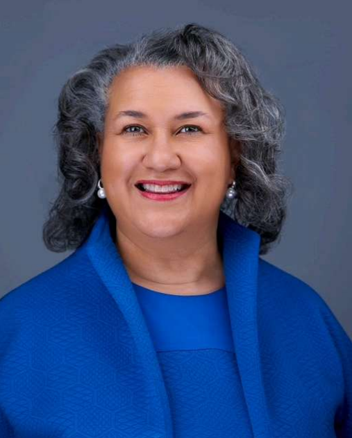 Headshot of Dr. Melissa Frank-Alston smiling, with shoulder-length gray hair, wearing a royal blue outfit and pearl earrings against a plain gray background.