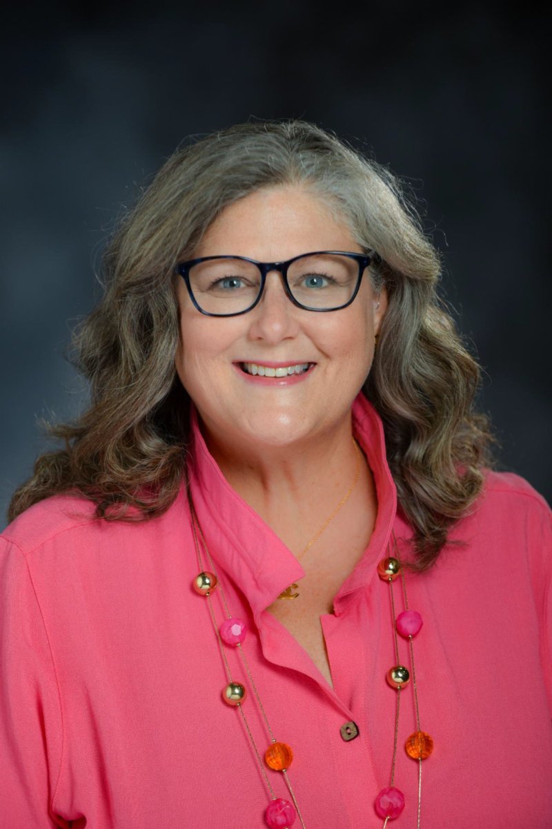 Smiling Caucasian woman wearing black glasses, a pink collared shirt and a necklace with pink, orange and silver balls posing for a photo
