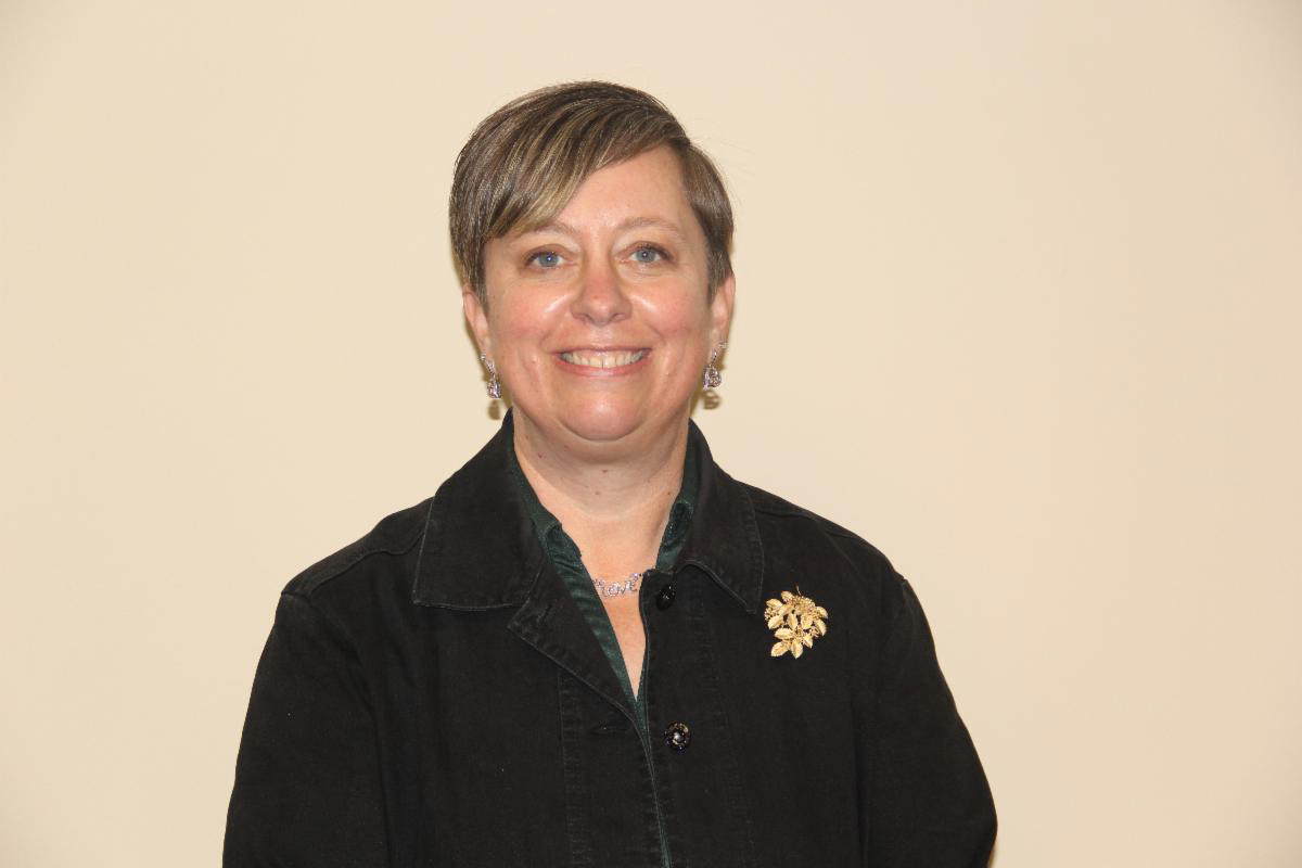 Caucasian woman with short blond and brown hair, wearing a black collared button down shirt, standing against a wall posing for a photo