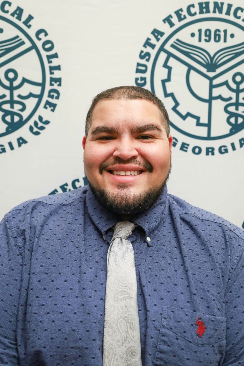 Colby Richardson, a Hispanic male, wears a light blue collared shirt with a darker blue pattern and silver grey tie while smiling at the camera against a background of the College Seal in Heritage Green on a white background.