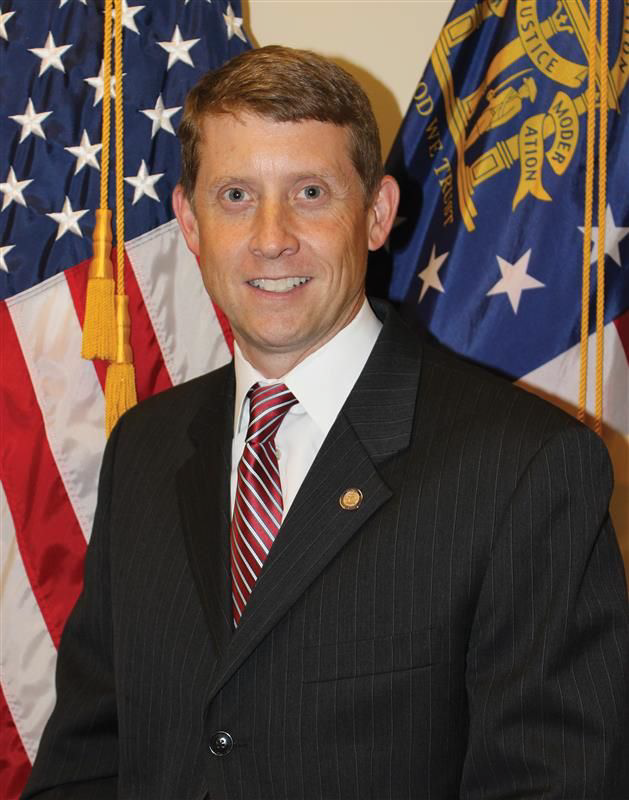 Mr. Greg Dozier, a Caucasian male, smiles at the camera wearing a black suit and white collared shirt with a red and white stirped tie. Pictured in front of the US flag and Georgia State flag.