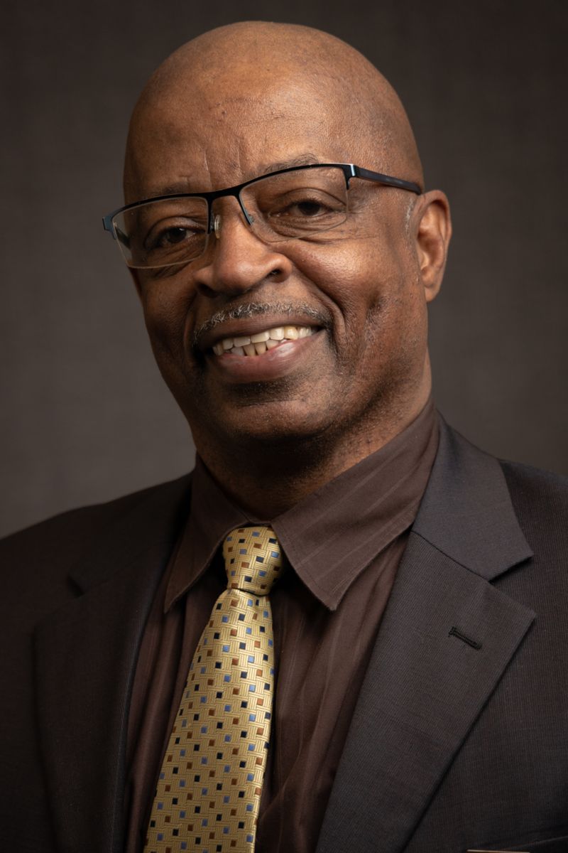 Harold Wright, an African American male wearing a grey suit jacket with a maroon collared shirt, a gold tie with black dots, and glasses.