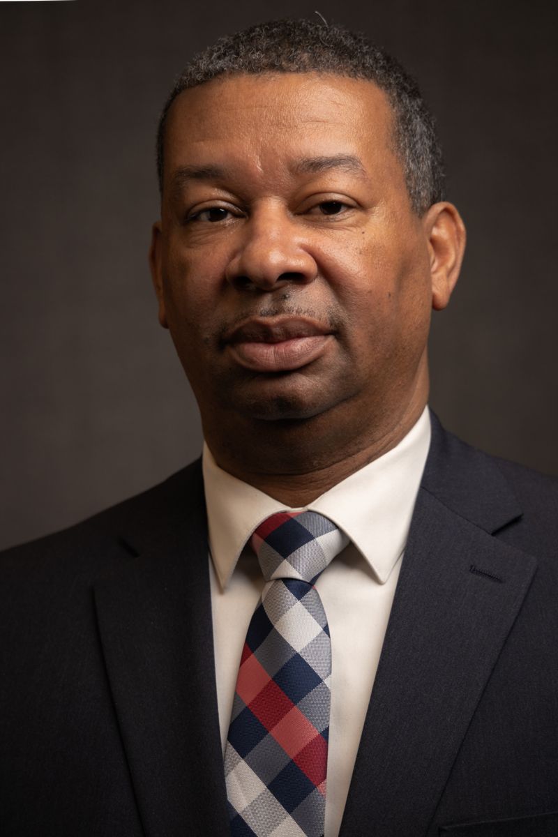 Dr. Howie Gunby, an African American male wearing a grey suit with a white collared shirt and a grey and white plaid tie with a single stripe of red.