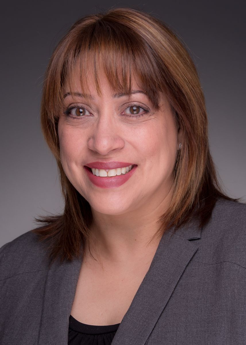 Migdaliz Berrios, a hispanic female with shoulder length brown hair is smiling and wearing a gray suit jacket with a black blouse.