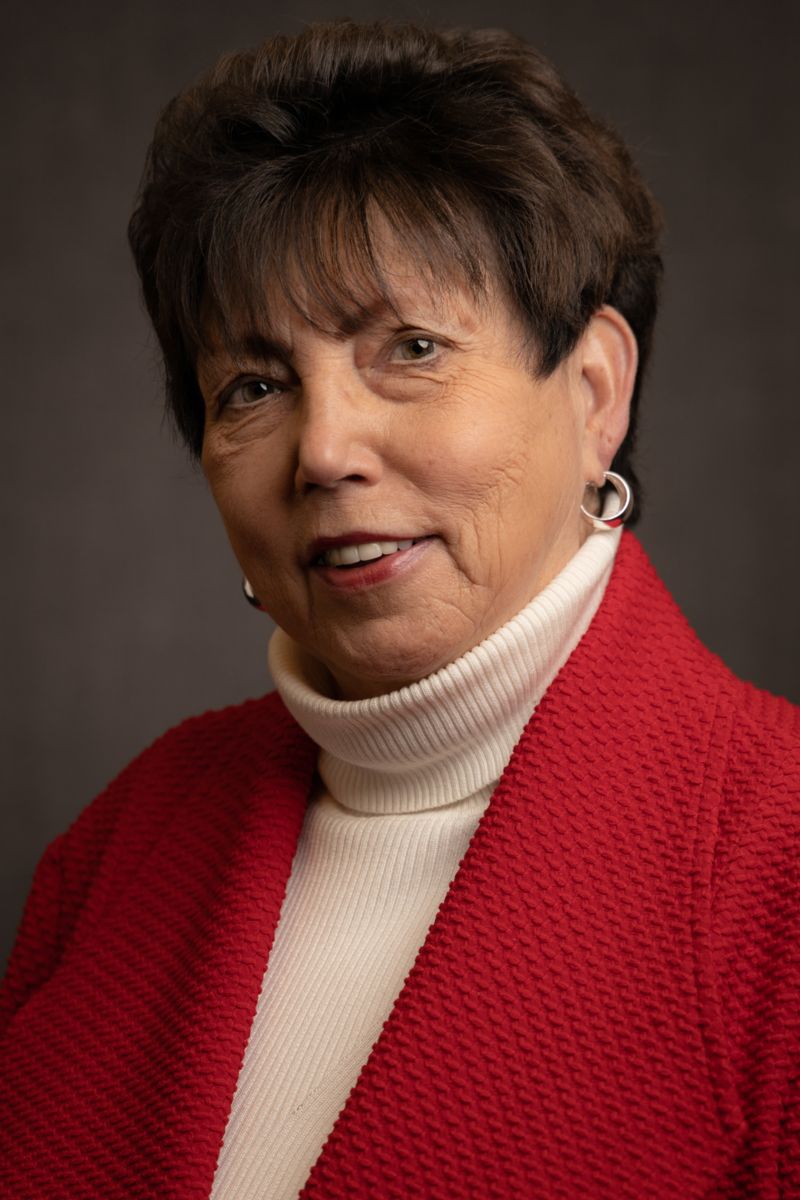 Pat Goodwin, a Caucasian female with short brown hair wearing gold earrings, a cream sweater, and a red cardigan.